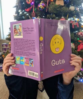 Child holding open a book to cover their face. The book is Guts by Raina Telgemeier