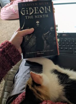A picture of a the book Gideon the Ninth being held in front of a laptop, with a kitten curled up in the foreground..