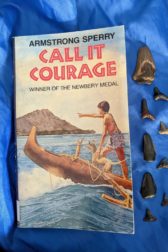 The book Call It Courage by Armstrong Sperry is on a blue cloth with fossilized shark teeth next to it
