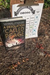 The book A Desolation Called Peace stands in front of a clipboard with a bingo sheet on it. The both are on dry ground with dead leaves and grasses.