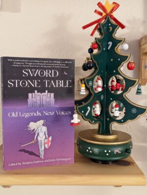 Picture of the book "Sword, Stone, Table: Old Legends, New Voice" is standing next to a miniature wooden Christmas tree hung with tiny ornaments.