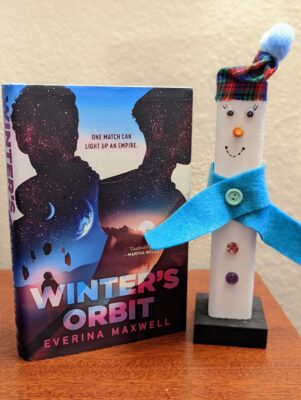 The book "Winter's Orbit" by Everina Maxwell stands next to a wooden snowman decoration.