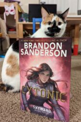 A calico cat sitting behind the book "Cytonic" by Brandon Sanderson.