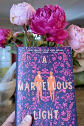 The book "A Marvellous Light" by Freya Marske held up in front of large light and dark pink peony flowers.
