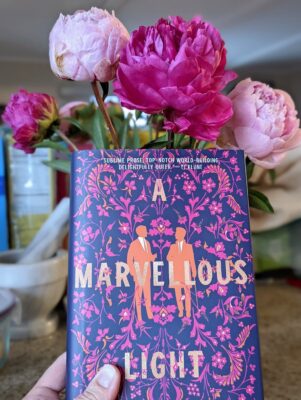 The book "A Marvellous Light" by Freya Marske held up in front of large light and dark pink peony flowers.