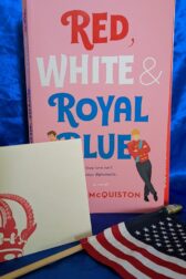 The book, "Red, White & Royal Blue" by Casey McQuiston standing behind a small American flag and a white notecard with an elaborate crown on it.