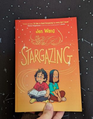 The book, "Stargazing" by Jen Wang held up in front of black fabric with little stars on it.