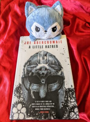 The book, "A Little Hatred" by Joe Abercrombie on a red satin background with a tiny, displeased looking plush wolf perched atop the book.