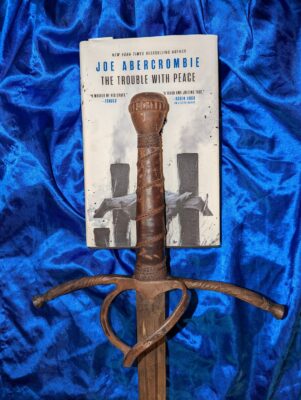 The book, "The Trouble with Peace" by Joe Abercrombie lying on a blue satin cloth with a rusty sword hilt lying on top of the book.