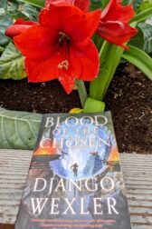 The book, "Blood of the Chosen" by Django Wexler lying in front of a red, blooming amaryllis flower.
