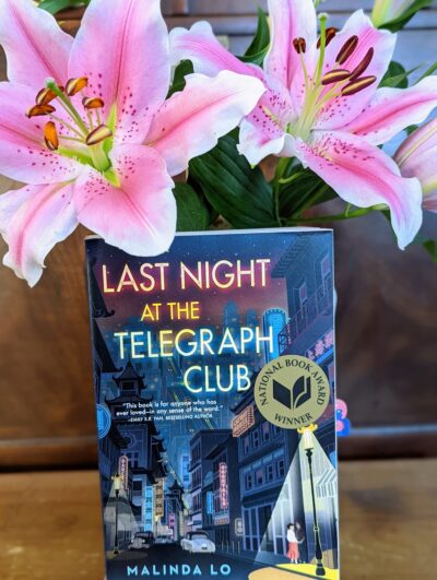 The book "Last Night at the Telegraph Club" by Malinda Lo in front of two blooming lilies.