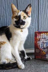 A calico cat sitting upright next to a the book, "Record of a Spaceborn Few" by Becky Chambers.