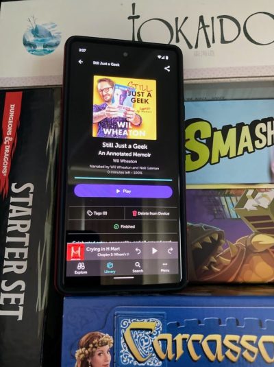 A cell phone showing an audiobook of "Still Just a Geek: An Annotated Memoir" by Wil Wheaton surrounded by board games.