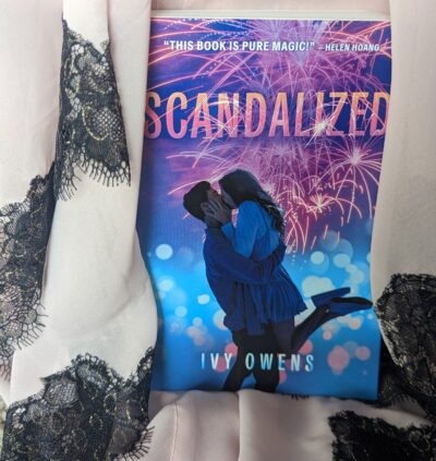 Pale pink fabric with black lace trim drapes around the book, "Scandalized" by Ivy Owens.