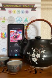 A small black tea pot with white flowers on it sits next to two tiny dark brown tea cups. Behind them is a cell phone showing the image of an audiobook. The audio book is "A Magic Steeped in Poison" by Judy I. Lin. In the background is a book bingo card.