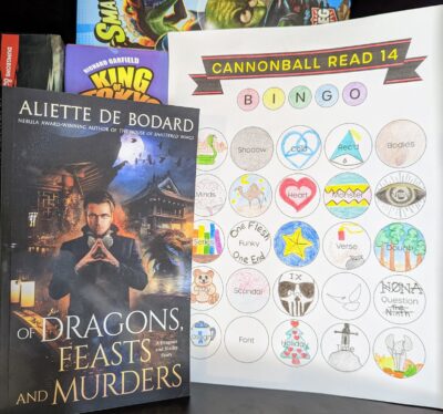 The book, "Of Dragons, Feasts, and Murder" by Aliette de Bodard standing next to an almost complete book bingo card.