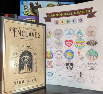 The book, "The Golden Enclaves" by Naomi Novik standing next to a book bingo card mostly filled out.