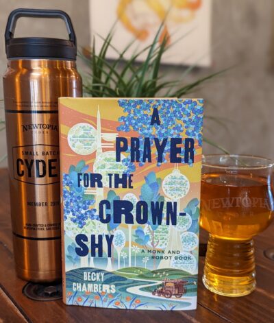 The book, "A Prayer for the Crown-Shy" by Becky Chambers standing next to a pint glass of hard cider and a bronze growler.
