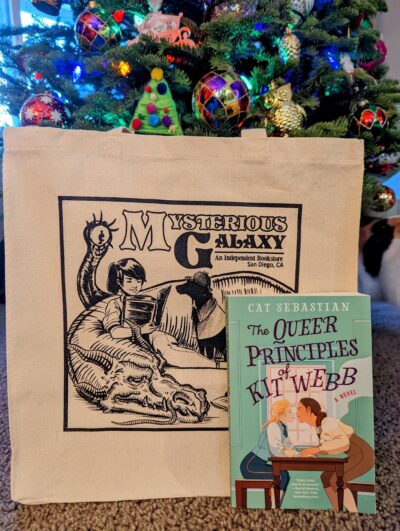 The book, "The Queer Principles of Kit Webb" by Cat Sebastian standing in front of a canvas tote bag for the bookstore, Mysterious Galaxy. In the background is a decorated Christmas tree.