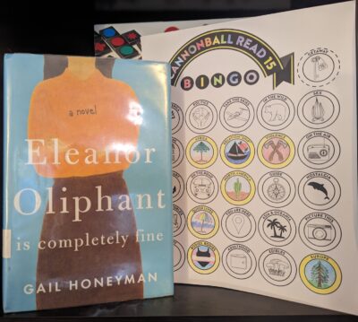 The book, "Eleanor Oliphant is Completely Fine", by Gail Honeyman, is standing next to a partially complete book bingo card.