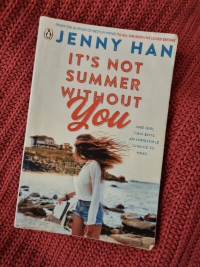 It's Not Summer Without You Cover. A girl on a beach in short denim shorts, with long hair, looking back towards a big beach house. 