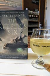 The book, "The Keeper's Six" by Kate Elliott standing next to a hard cider from Serpentine Cider.