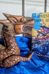 The book, "The Adventures of Amina al-Sirafi" by Shannon Chakraborty standing next to a wooden dragon-like sculpture, resting on shiny blue fabric giving the impression of water.