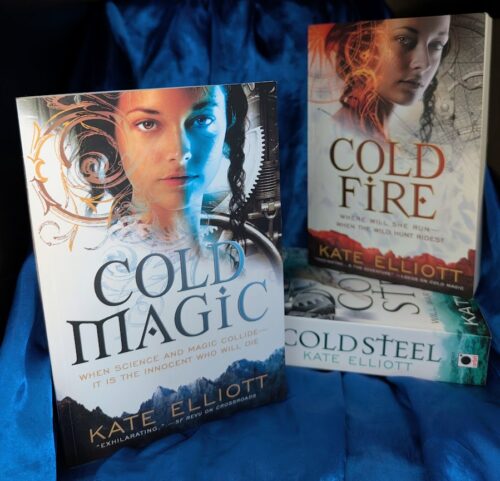The books, "Cold Magic", "Cold Fire", and "Cold Steel" by Kate Daniels artfully arranged on blue satin cloth.
