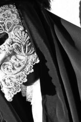 Black and white photo of a sinister figure wearing black robes and an ornate white mask.