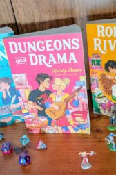 Three books, "Dungeons and Drama", "Dragons and Dating", "Rolls and Rivalries", by Kristy Boyce are standing in a row on a bookshelf. In front of them are a collection of dice in multiples shapes and miniature figures for playing Dungeons and Dragons.