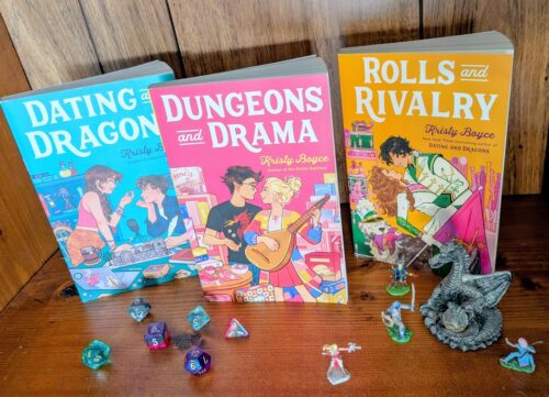 Three books, "Dungeons and Drama", "Dragons and Dating", "Rolls and Rivalries", by Kristy Boyce are standing in a row on a bookshelf. In front of them are a collection of dice in multiples shapes and miniature figures for playing Dungeons and Dragons.